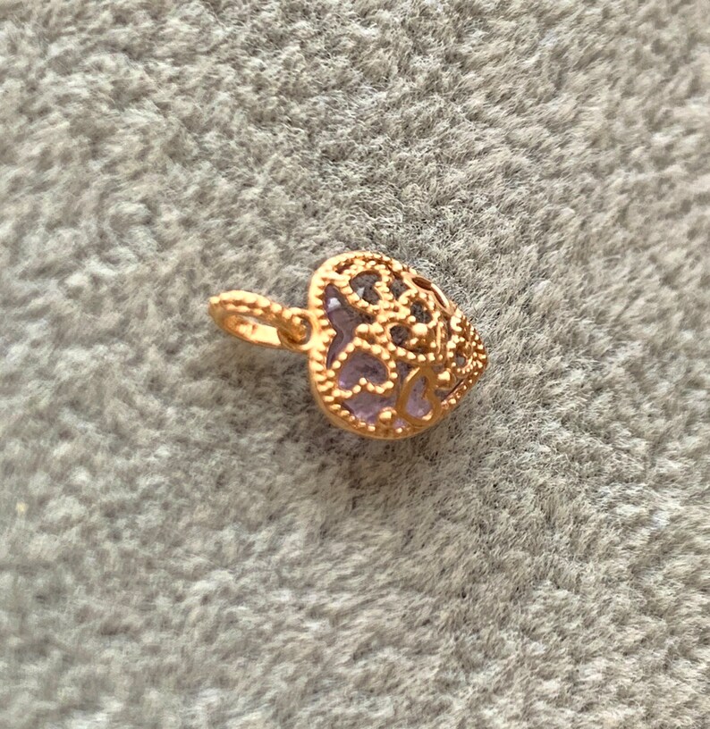 K&ouml;nnte beinhalten: Ein goldfarbener herzf&ouml;rmiger Anh&auml;nger mit einem filigranen Design und einem violetten Stein in der Mitte.