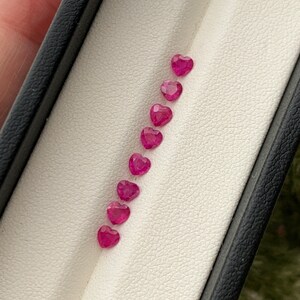 May include: A row of ten heart-shaped pink gemstones in a white display case.