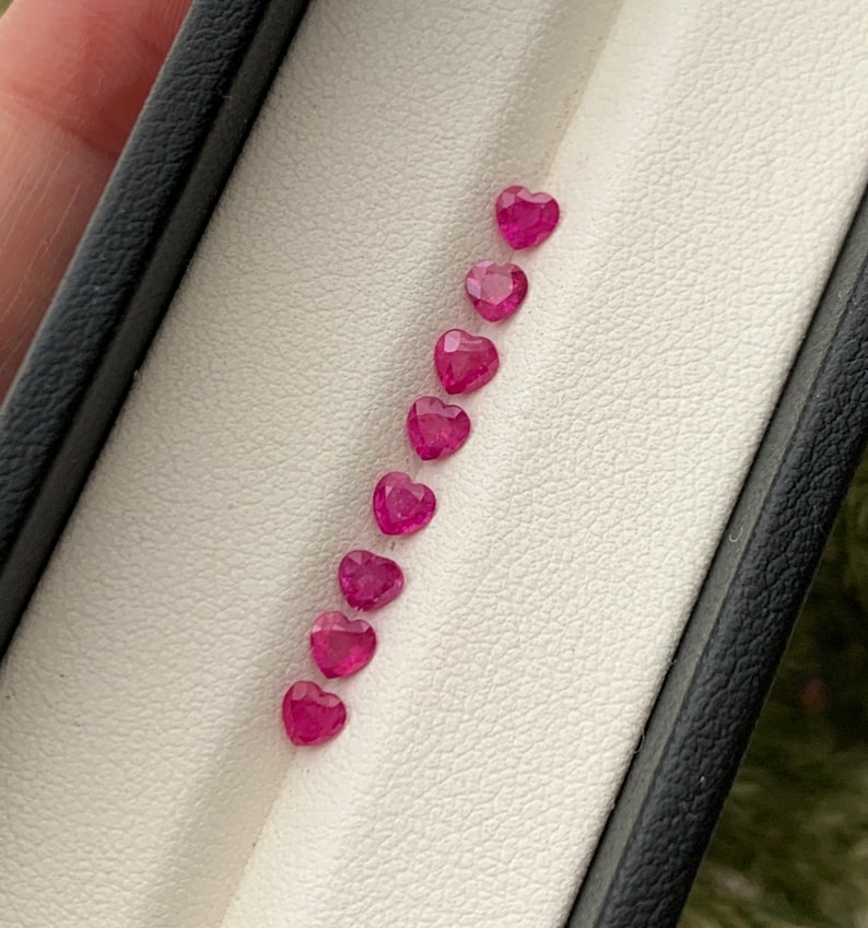 May include: A row of ten heart-shaped pink gemstones in a white display case.