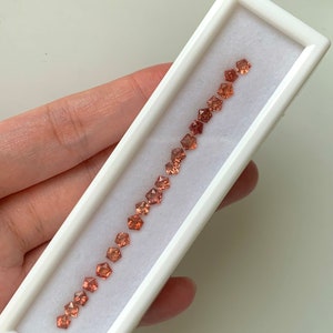 May include: A white box with a row of 18 small, faceted, orange-brown gemstones. The gemstones are arranged in a line in the box.