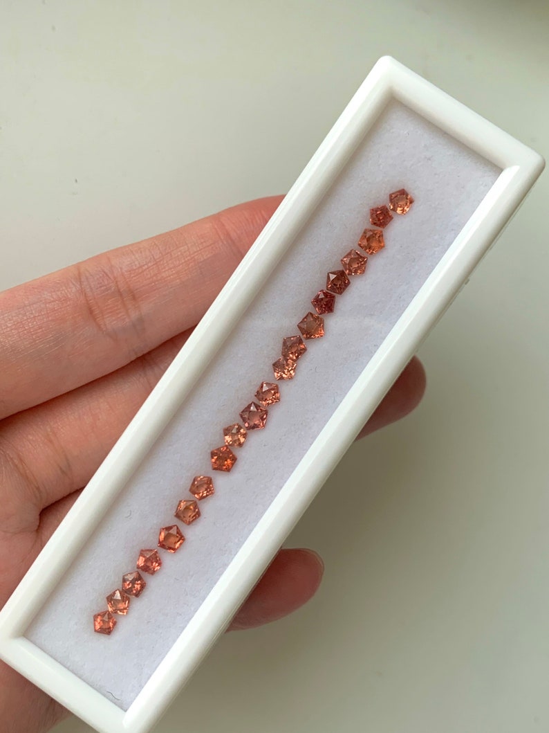 May include: A white box with a row of 18 small, faceted, orange-brown gemstones. The gemstones are arranged in a line in the box.