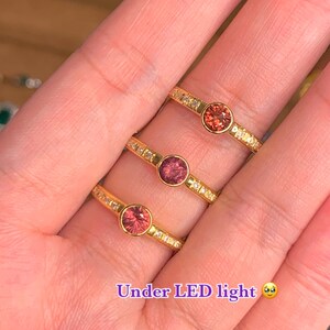 May include: Three gold rings with a single round gemstone in each. The gemstones are different shades of pink and red. The rings are set with small clear stones around the center stone. The caption reads "Under LED light".