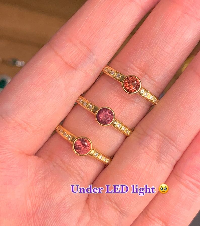 May include: Three gold rings with a single round gemstone in each. The gemstones are different shades of pink and red. The rings are set with small clear stones around the center stone. The caption reads "Under LED light".