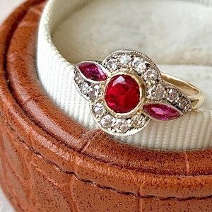Könnte beinhalten: Ein Goldring mit einem zentralen, rund geschliffenen roten Edelstein, der von einem Kranz aus kleinen klaren Steinen umgeben ist. Zwei marquise-geschliffene rote Steine befinden sich an den Seiten, flankiert von weiteren klaren Steinen.