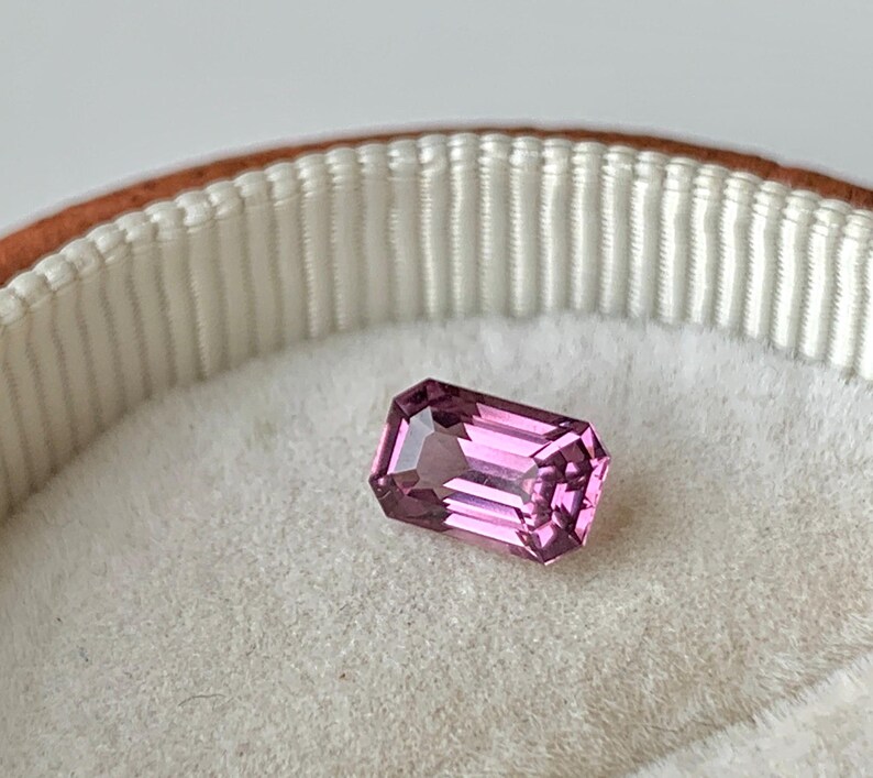 May include: A pink gemstone with a rectangular emerald cut, displayed in a white velvet ring box.