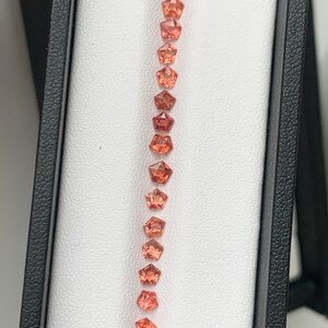 May include: A row of 18 orange and pink gemstones in a white box. The gemstones are all octagonal in shape and are arranged in a vertical line.
