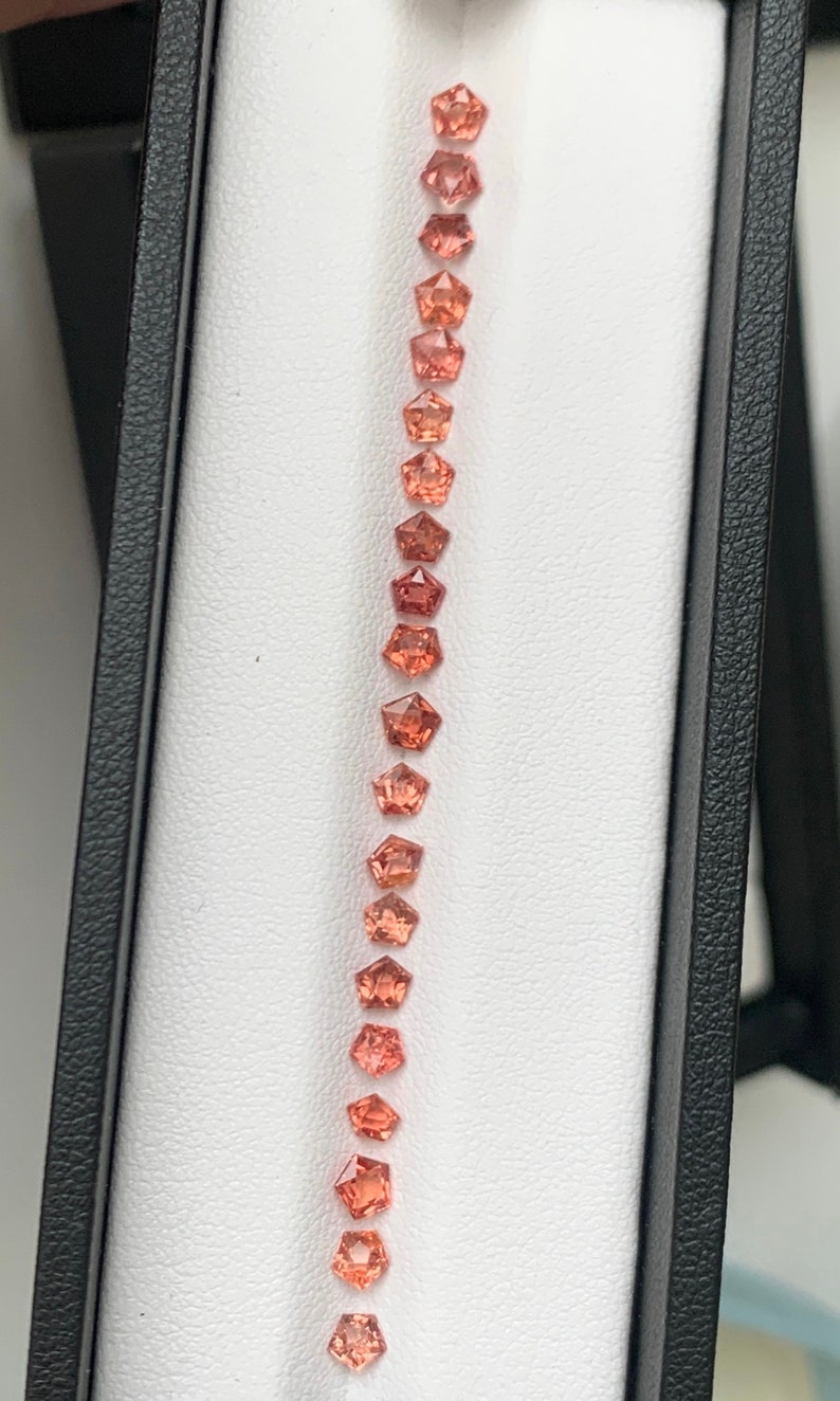 May include: A row of 18 orange and pink gemstones in a white box. The gemstones are all octagonal in shape and are arranged in a vertical line.