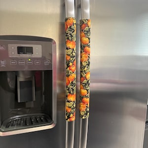 Puede incluir: Fundas para manijas de refrigerador con un estampado de calabazas otoñal. Las fundas presentan un fondo negro con calabazas naranjas, hojas verdes y enredaderas. Están diseñadas para adaptarse a las manijas de acero inoxidable de un refrigerador.