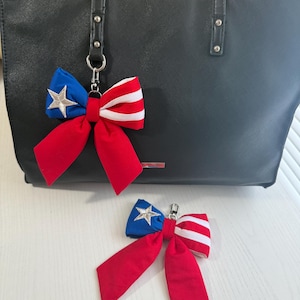 Puede incluir: Un bolso tote negro con un llavero de lazo rojo, blanco y azul. El lazo presenta una sección azul con una estrella plateada, una sección roja y una sección a rayas blancas y rojas. Un segundo lazo está en la superficie.