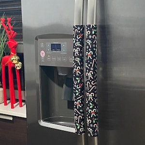 Könnte beinhalten: Zwei schwarze Kühlschrank-Griffbezüge mit einem Weihnachts-Zuckerstangen- und Stechpalmenmuster. Die Bezüge sind aus Stoff und sollen die Griffe vor Abnutzung schützen.