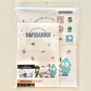 May include: A clear plastic package containing a letter set with the text "HAPIDANBUI". The set includes letter sheets and envelopes featuring cartoon characters and food illustrations. The text "GOOD FOOD, GREAT COMPANY" is also visible.