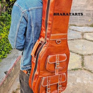 Könnte beinhalten: Eine braune Gitarrentasche aus Leder mit mehreren Taschen und Schnallen. Die Tasche hat einen Trageriemen für den Transport auf dem Rücken. Der Text "BHARATARTS" ist auf die Tasche gedruckt.