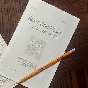 May include: A white workbook titled Restoring Peace to your mornings with a circular logo, a yellow Staedtler pencil, an open gardening textbook with green accents, and a slice of bread in a plastic bag on a dark brown wooden surface.