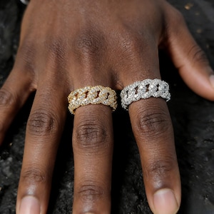 May include: Two rings with a chain link design, one gold-toned and one silver-toned, both encrusted with small, clear gemstones. The rings are displayed on a dark surface, showcasing their intricate details and sparkling appearance. The rings are a jewelry item.