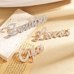 Puede incluir: Tres broches con nombres de pedrería en tonos plateados y dorados. Los nombres "Lenba", "Lucas" y "Cyfa" están escritos en cursiva y están incrustados con cristales brillantes. Estos broches son perfectos para añadir un toque personal a cualquier atuendo.