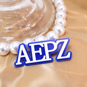 Könnte beinhalten: Ein blau-weißer AEPZ-Anhänger mit weißem Rand, vor goldenem Satinstoff. Eine Kette aus weißen Perlen und ein klarer Glasgegenstand sind ebenfalls im Bild. Der Anhänger ist der Blickfang.