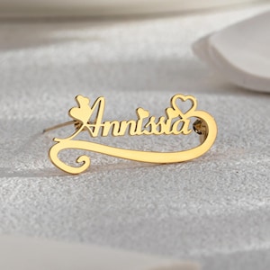 May include: A gold-coloured brooch with the name "Annissia" in a cursive font. The brooch features heart and floral accents. The design is a personalised jewellery item, likely a pin or a pendant.