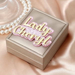 Può includere: Un portagioie quadrato beige con una targhetta dorata "Lady Cheryl". La targhetta ha uno sfondo glitterato rosa e scritte dorate. Una collana di perle è visibile sullo sfondo.