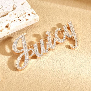 Könnte beinhalten: Eine goldfarbene Brosche mit dem Wort "Juicy" in Schreibschrift. Die Buchstaben sind mit funkelnden, klaren Kristallen besetzt. Die Brosche ist ein Modeaccessoire.
