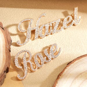 May include: Two gold-toned brooches, one reading "Hazel" and the other "Rose", are encrusted with sparkling crystals. The brooches are in a cursive font and are set against a neutral background, suggesting a jewelry or accessory theme.