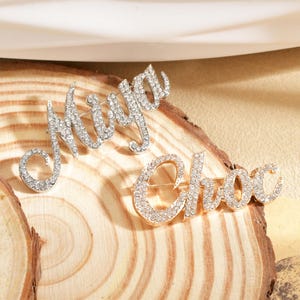 Op de afbeelding: Twee met strass bezette broches met de letters "Miya" en "Choc". De "Miya" broche is zilverkleurig, terwijl de "Choc" broche goudkleurig is. Beide worden op een houten oppervlak gepresenteerd.