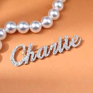 以下が含まれることがあります： 「Charlie」の文字が筆記体で書かれた、クリアクリスタルがちりばめられたシルバーカラーのブローチ。暖かくニュートラルな背景に配置され、画像上部には白い真珠のネックレスが添えられています。