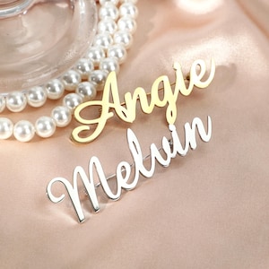 Puede incluir: Dos broches con nombres, uno dorado y otro plateado, se muestran sobre una superficie rosa. El broche dorado deletrea "Angie" en cursiva. El broche plateado deletrea "Melvin" en un estilo cursivo similar. Un collar de perlas está en el fondo.