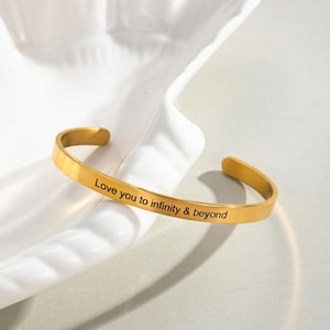 May include: A gold-colored cuff bracelet with the inscription "Love you to infinity & beyond." The open-ended bracelet is resting on a white, sculpted surface. The bracelet has a smooth, polished finish and a simple, elegant design.