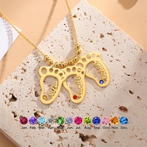 May include: A gold-colored necklace featuring three pendant footprints, each with a name engraved. The pendants are adorned with colorful gemstones. The necklace is displayed on a textured surface with a selection of birthstone options.