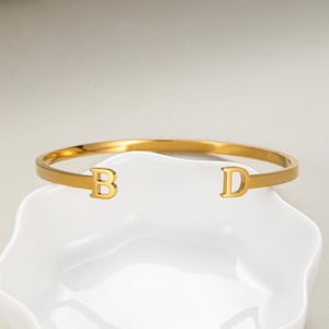 May include: A gold-coloured bangle bracelet featuring the letters "B" and "D" at each end. The bracelet is displayed on a white, scalloped-edge dish. The bracelet has a smooth, polished finish and a simple, elegant design.