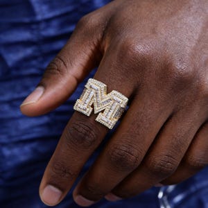 以下が含まれることがあります： 小さな輝く石がちりばめられた、金色の「M」の文字が特徴のリング。リングの複雑なデザインと石の輝きが、暗い背景の中で際立っています。大胆なジュエリーです。