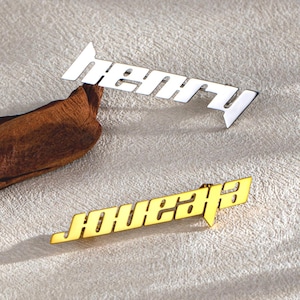 May include: Two metal bar pins, one silver and one gold, each with a name in a stylized font. The silver pin reads "Henry" and the gold pin reads "Eleanor". Both pins are on a neutral-toned surface.
