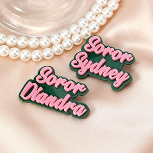 以下が含まれることがあります： 「Soror Diandra」と「Soror Sydney」という言葉が筆記体で書かれた、緑とピンクの名札が2つ。名札はサテンの表面にあり、背景には真珠のネックレスがあります。