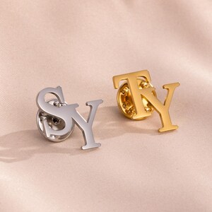 May include: Two initial pins, one silver and one gold, are displayed on a soft, peach-colored surface. The silver pin spells "SY", while the gold pin spells "TY". Each pin has a pin back.