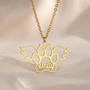 May include: A gold-colored necklace featuring a paw print design with angel wings and the name "Emily" in a script font. The pendant hangs from a delicate chain, creating a personalized pet memorial.