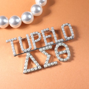 May include: A silver-toned brooch spelling "TUPELO" with pearl accents, along with Greek letters. A strand of large, white pearls is in the background. The brooch is on an orange surface.