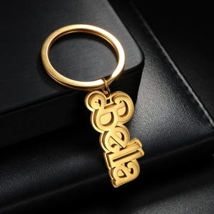 May include: A gold-coloured keychain featuring the name "Bella" in a decorative font. The keychain has a large circular ring and a smaller ring connecting to the name pendant. The background is a dark, textured surface.