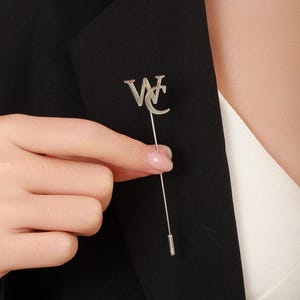 May include: A silver-colored lapel pin with the letters "WC" in a stylized design. The pin is held by a hand against a black blazer, with a white shirt visible underneath. The pin is a long, thin stick pin.