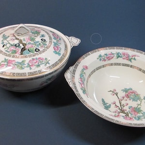 May include: A set of two white ceramic bowls with a floral design. The bowls have a gold trim and feature a pink and green floral pattern with a tree branch and flowers.
