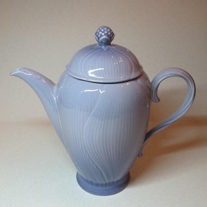 Puede incluir: Una tetera de cerámica azul claro con tapa y pomo en la parte superior. La tetera tiene un diseño texturizado y un cuerpo redondeado.