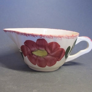 Op de afbeelding: Witte keramische sauskom met een roze bloem en groene bladeren ontwerp. De kom heeft een gegolfde rand en een handvat.