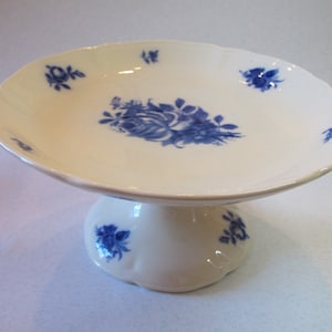 May include: A white cake stand with a blue floral design. The stand has a scalloped edge and a pedestal base.