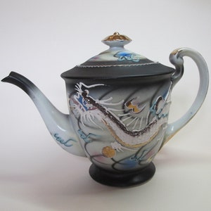 May include: A white teapot with a black dragon design and gold accents. The teapot has a lid with a gold knob and a handle on the side. The dragon is depicted in a swirling pattern with blue, gold, and white accents.