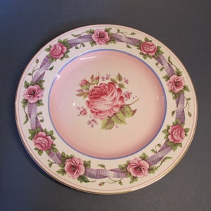 Peut inclure: Une assiette en céramique rose et blanche avec un motif floral. L'assiette a un large bord avec un ruban violet et des roses roses. Le centre de l'assiette présente une grande rose rose avec des feuilles vertes.