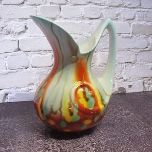 Puede incluir: Una jarra de cerámica colorida con un diseño único. La jarra tiene un patrón de remolino de color verde claro, naranja, amarillo y marrón. La jarra tiene una boca ancha y un asa.