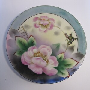 May include: A vintage porcelain plate with a blue rim and a floral design. The plate features two pink roses with green leaves and a small bird perched on a branch.