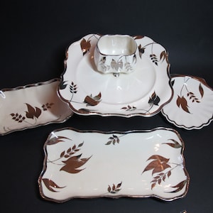 Peut inclure: Un ensemble de quatre plats de service en céramique blanche avec des accents en feuilles d'argent. Les plats présentent un motif répétitif de feuilles et d'épis de blé. L'ensemble comprend un plat carré avec un petit bol au centre, un plat rectangulaire et deux petits plats ronds.