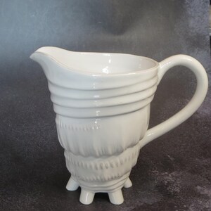 May include: White ceramic pitcher with a curved spout and a large handle. The body features horizontal ridged detailing and a textured lower section, supported by three small feet. A classic serving piece.