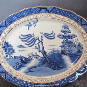 May include: Oval blue and white ceramic serving platter with a scalloped edge and gold trim. The platter features a detailed landscape scene with a bridge, pagoda, and birds in flight. The border has a geometric pattern.