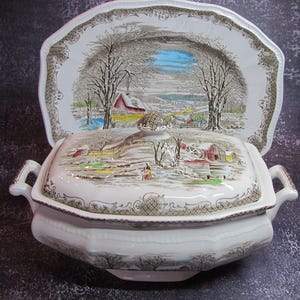 May include: A white ceramic serving dish and matching platter, both decorated with a winter landscape. The dish has a lid with a decorative knob and handles. The platter is scalloped and features the same winter scene.
